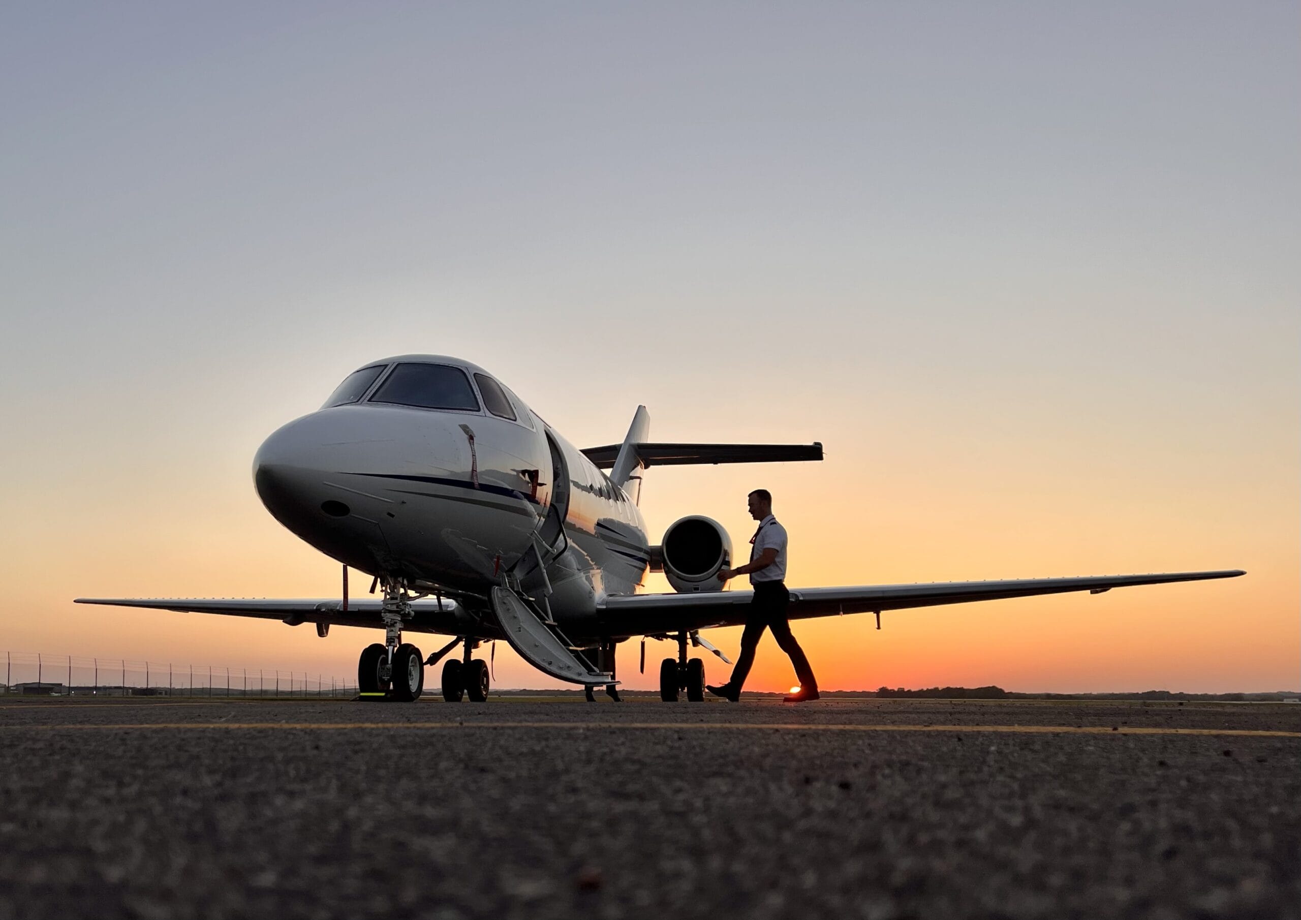 A pilot walks towards a private jet at sunset, with vibrant orange and purple skies in the background.
