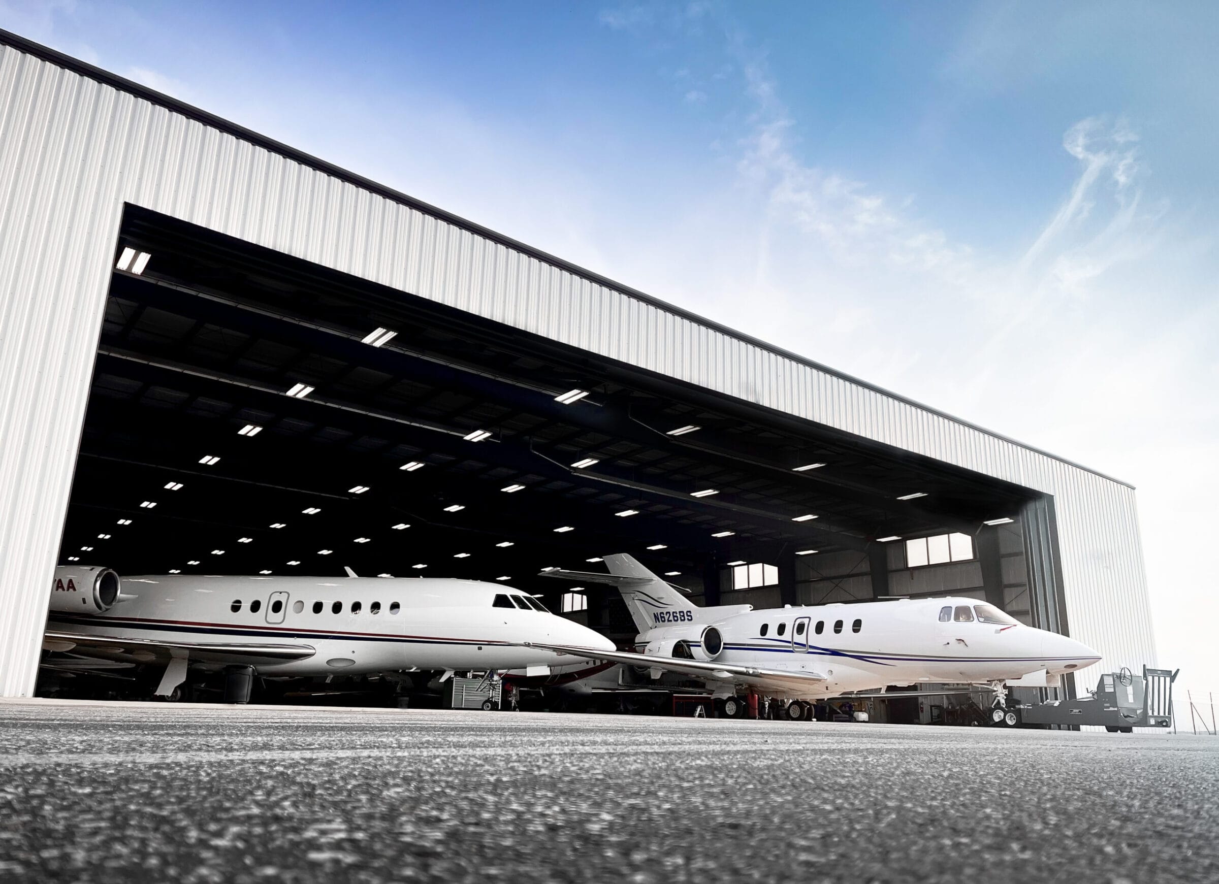 Two private jets parked inside a large hangar, with bright overhead lights and a clear blue sky visible outside.