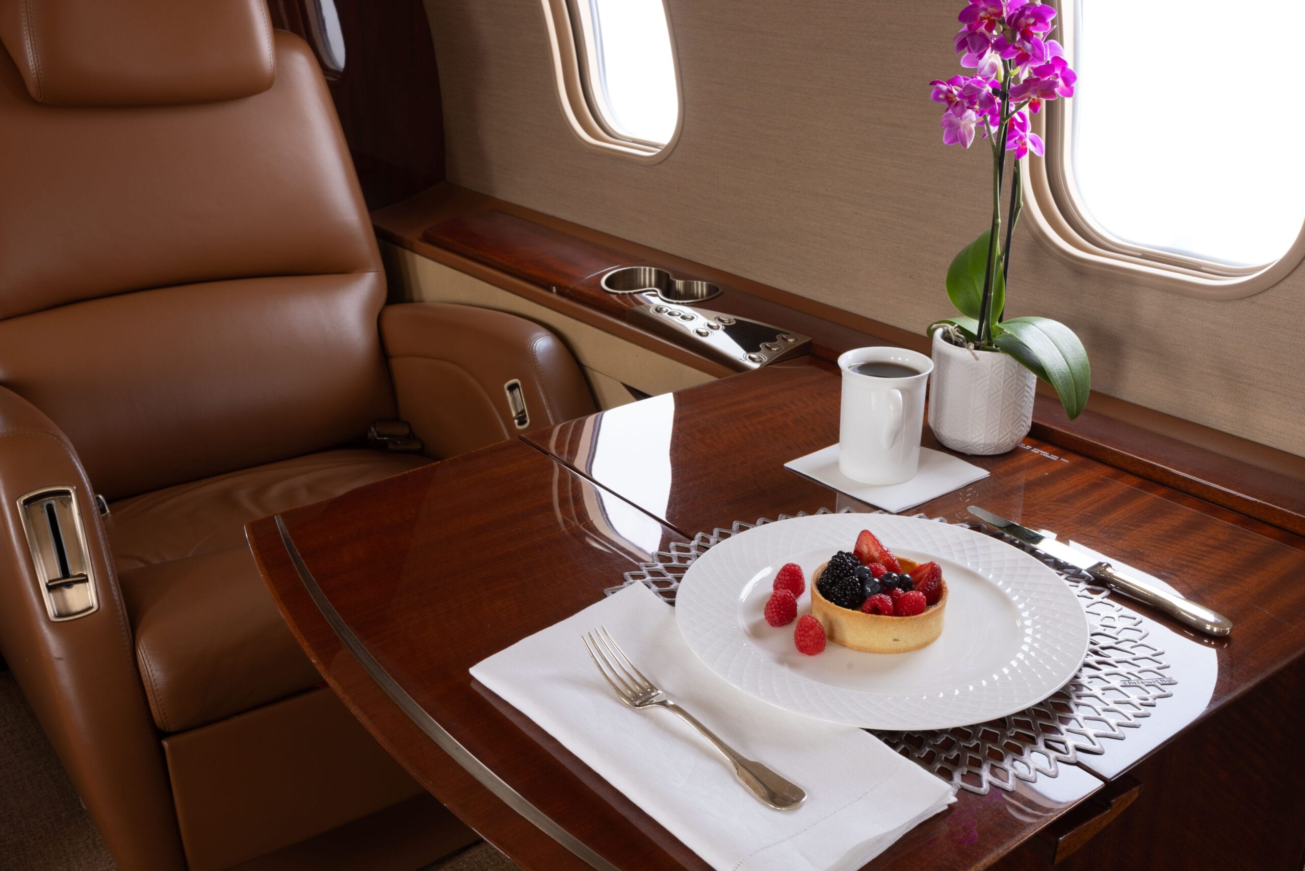 Luxurious airplane interior featuring a leather seat, a gourmet fruit tart, coffee, and a potted orchid on a polished table.