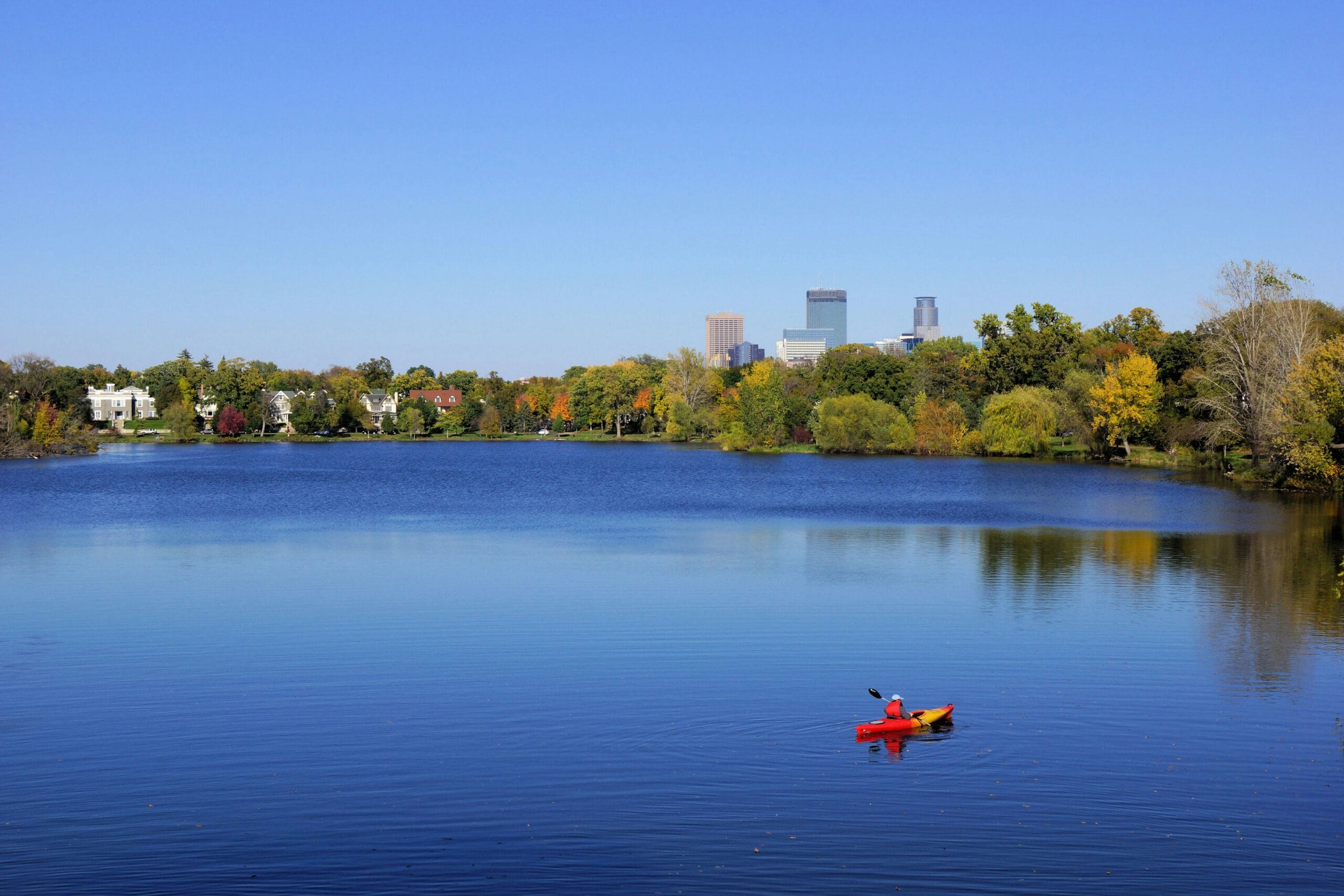 Kayaker on a blue lake with autumn trees and city skyline in background.