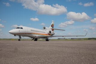 White private jet with rainbow stripe and logo on tail, parked on tarmac under cloudy sky.