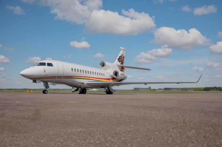 White private jet with rainbow stripe and logo on tail, parked on tarmac under cloudy sky.
