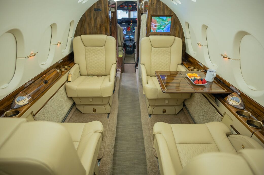 Luxurious private jet interior with cream leather seats, wood accents, and a tray of fruit.