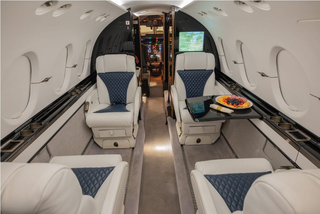 Luxury jet interior with white and blue leather seats, a tray of fruit, and a cockpit view.